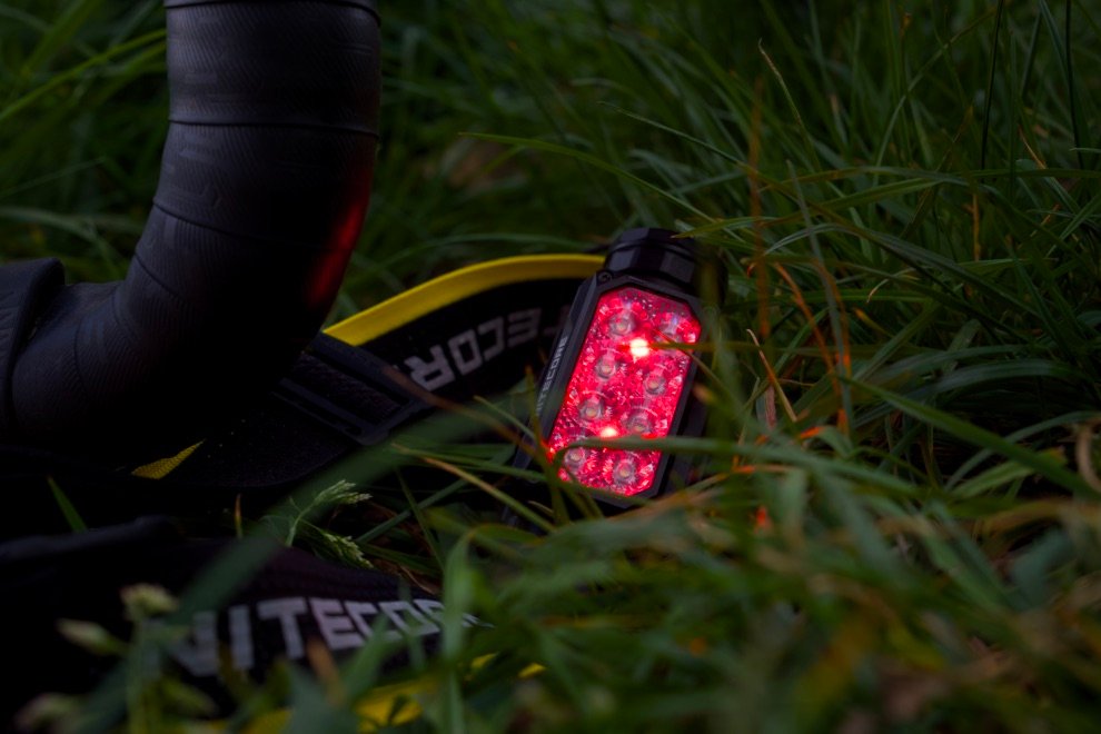 Test & Avis Nitecore HC65 UHE : La lampe frontale ultime pour le bikepacking ? 6 Lampe frontale Nitecore HC65 UHE en mode rouge posée dans l’herbe, idéale pour préserver la vision nocturne en bivouac