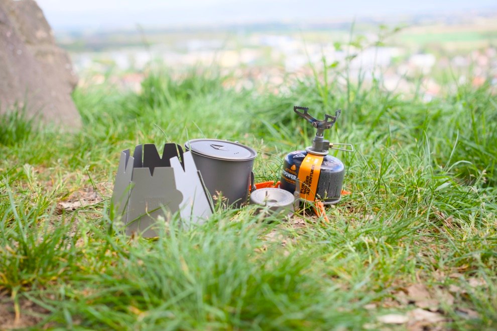Matériel de bivouac posé dans l’herbe : réchaud à gaz, popote en aluminium, coupe-vent et cartouche de gaz, prêt à l’usage en extérieur