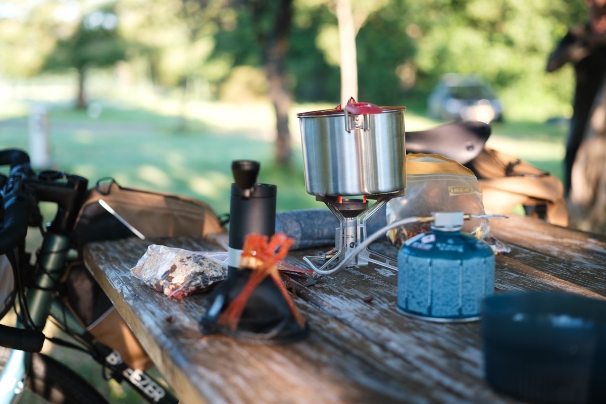 Préparation d’un petit déjeuner en bikepacking sur une table de pique-nique avec réchaud à gaz, popote en inox et sachet de muesli