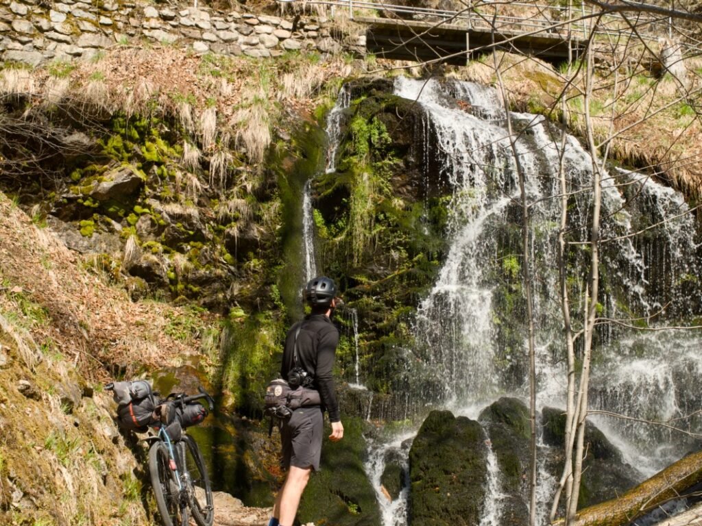 Traversée de la Forêt-Noire en gravel : 544 km en 5 jours, du pneu éclaté à la tente gelée 12 Cycliste en tenue noire et casque devant une cascade tombant sur des rochers moussus, son gravel chargé de sacoches posé sur le chemin à gauche