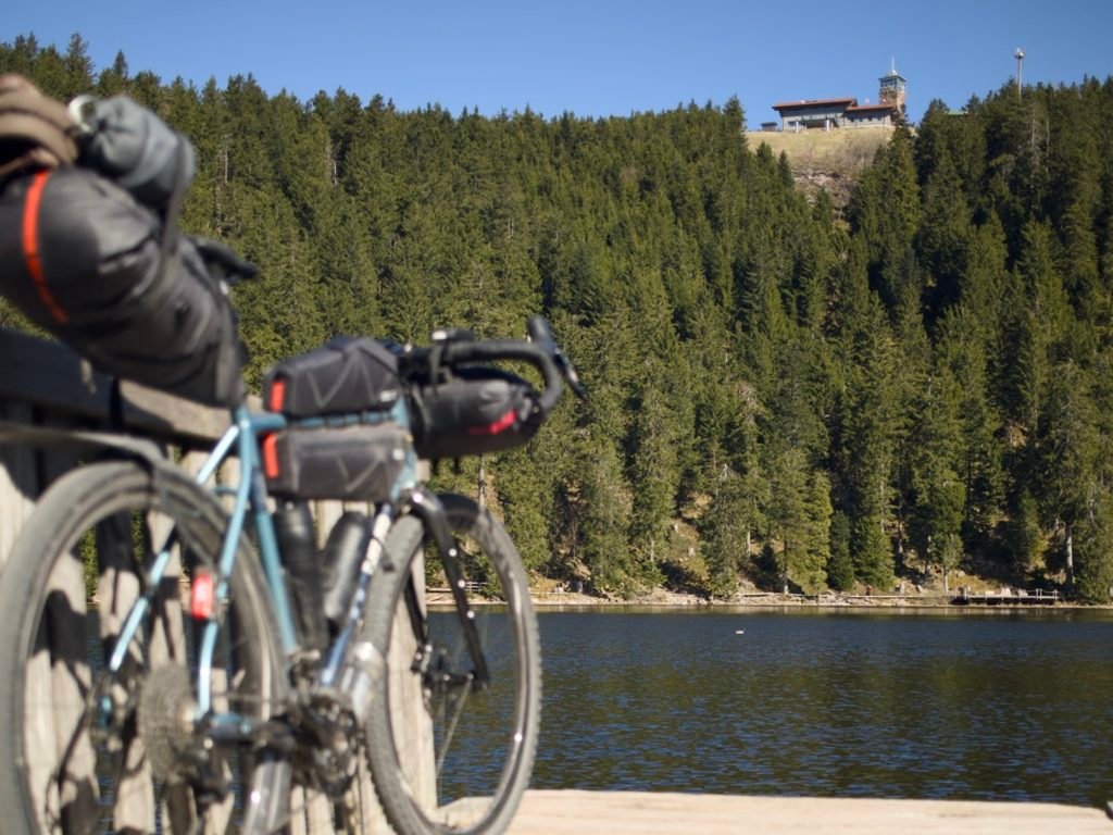 Traversée de la Forêt-Noire en gravel : 544 km en 5 jours, du pneu éclaté à la tente gelée 9 Vélo gravel bleu chargé de sacoches de bikepacking, appuyé sur un ponton en bois face au Mummelsee, un lac entouré de sapins en Forêt-Noire