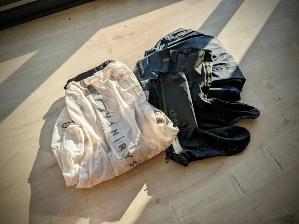 Quels vêtements emporter en bikepacking ? 6 Équipement de pluie pour le bikepacking posé sur un parquet : veste coupe-pluie Van Rysel blanche translucide, couvre-chaussures DHB noirs et vêtement de pluie gris foncé.