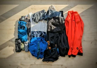 Quels vêtements emporter en bikepacking ? 5 Vêtements de bikepacking étalés sur un parquet, vus de dessus : veste thermique orange, maillot bleu, collant long, sous-couches, chaussettes en mérinos et couvre-chaussures.