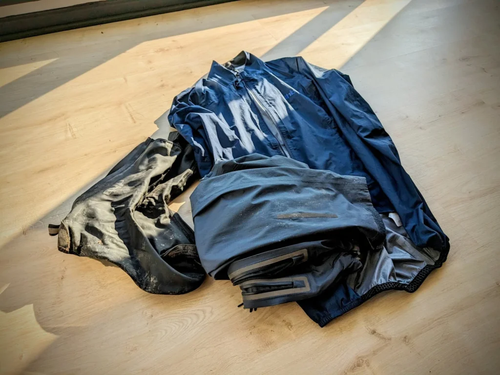 Quels vêtements emporter en bikepacking ? 7 Vêtements de pluie pour le bikepacking posés sur un parquet : veste de pluie bleu marine, surpantalon gris et couvre-chaussures tachés de boue.