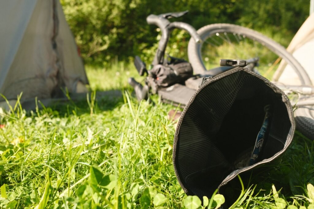 Sacoche ouverte posée dans l'herbe au bivouac, avec en arrière-plan un vélo couché équipé d'un compteur GPS sur le cintre, entre deux tentes.